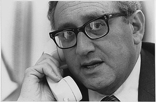 Henry Kissinger wearing glasses and a suit is engaged in a phone conversation, appearing focused and professional.