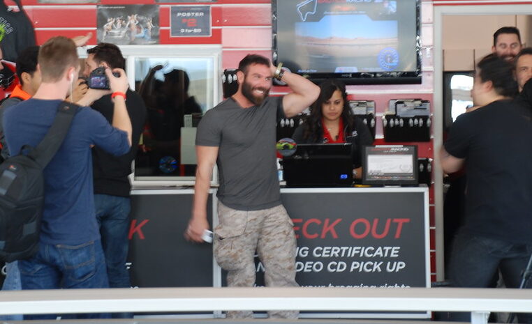 Dan Bilzerian stands near a counter smiling, with people around him taking photos. The setting appears to be an indoor event or venue.